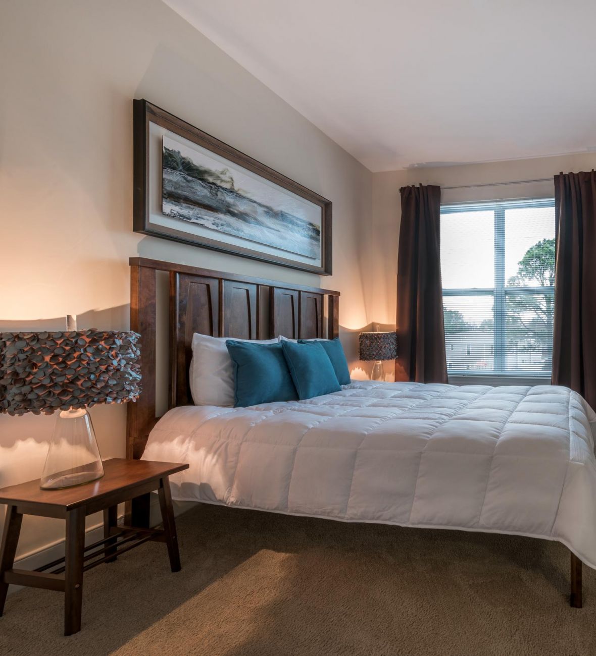 Infinity-at-Centerville-Crossing Modern bedroom with a wooden bed, blue pillows, art on the walls, and light from a window.