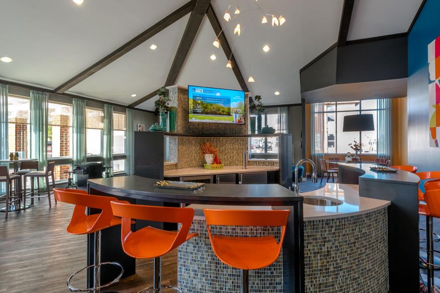 Infinity at Centerville Crossing Modern lounge with orange bar stools, a curved counter, wall art, TV, and large windows letting in natural light.
