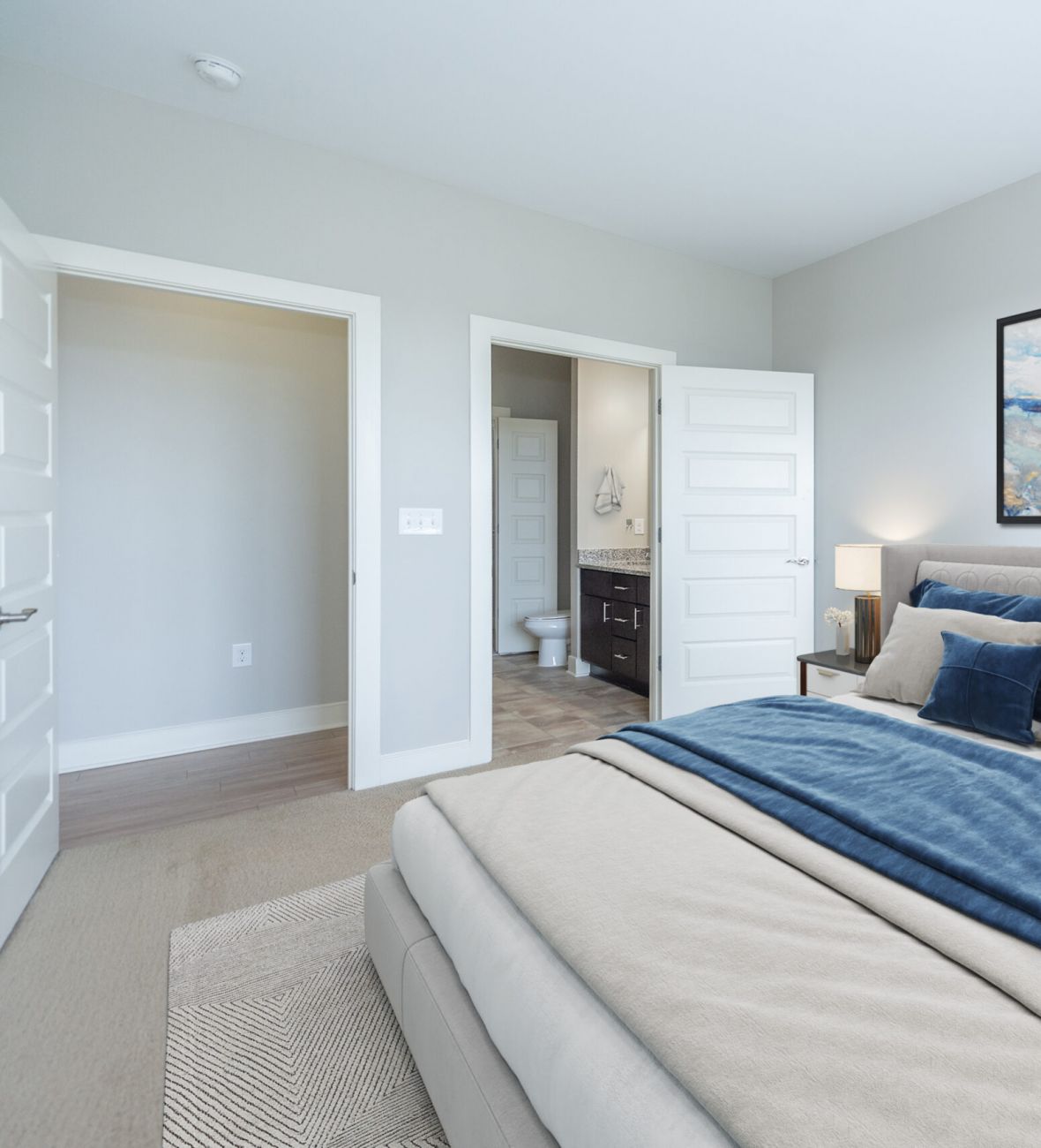 Infinity apartment homes with Modern bedroom with blue and beige bedding, abstract wall art, and an open door leading to an en-suite bathroom.
