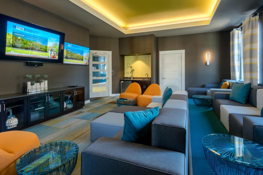 Infinity at Centerville Crossing Modern lounge with gray sofas, orange chairs, blue pillows, wall-mounted TVs, and accent lighting.