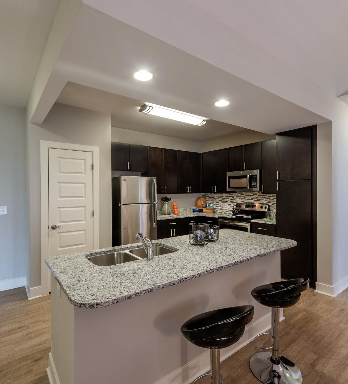 Infinity-at-Centerville-Crossing Modern kitchen with dark cabinets, granite island, and two black barstools on wood flooring.