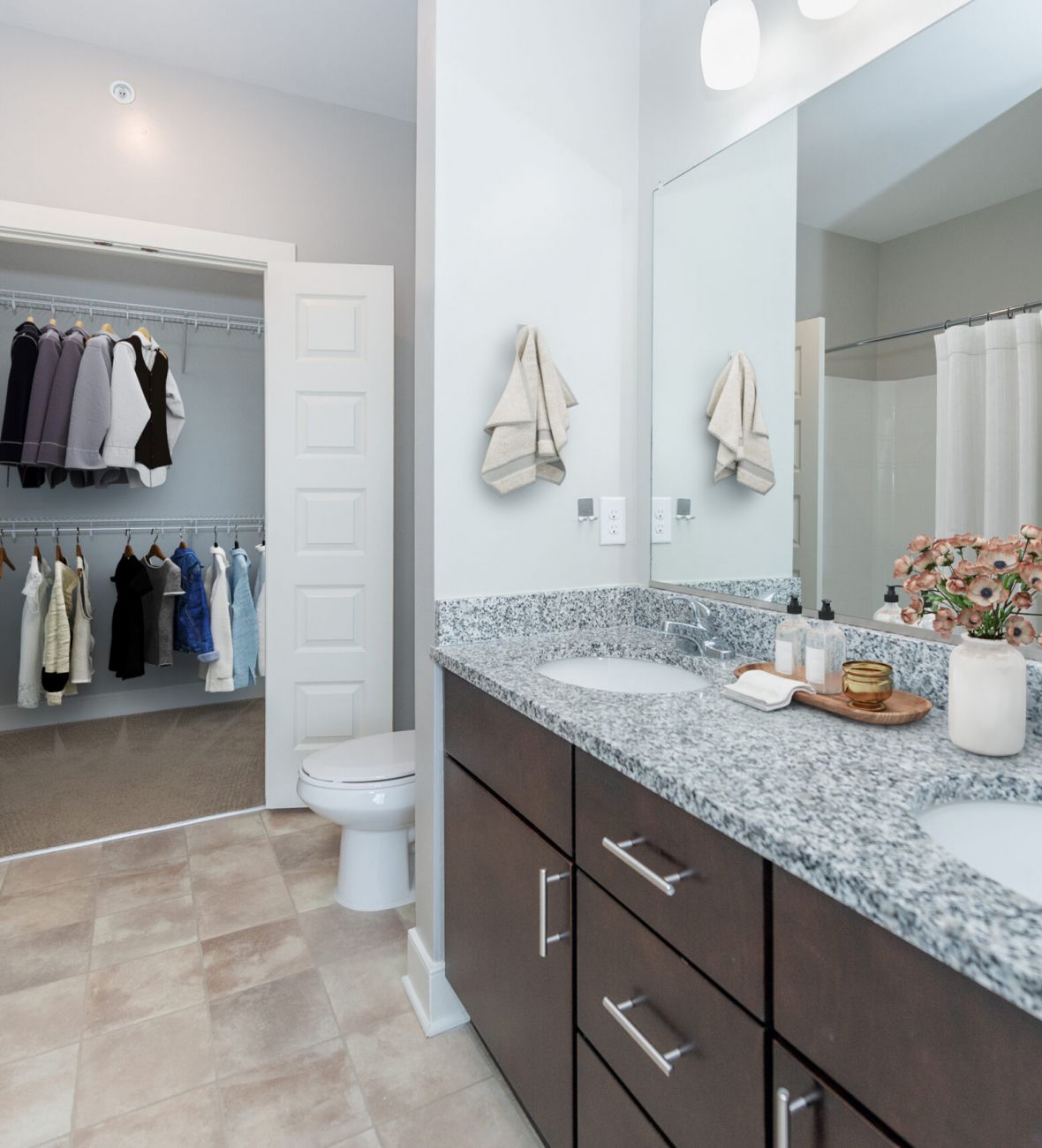 Infinity apartment homes with Modern bathroom with double sink vanity, flowers, and a view into a walk-in closet with hanging clothes.
