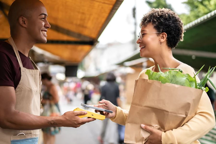 Infinity at Centerville Crossing Two people smile as one pays with a phone at an outdoor market while holding a paper bag of groceries.