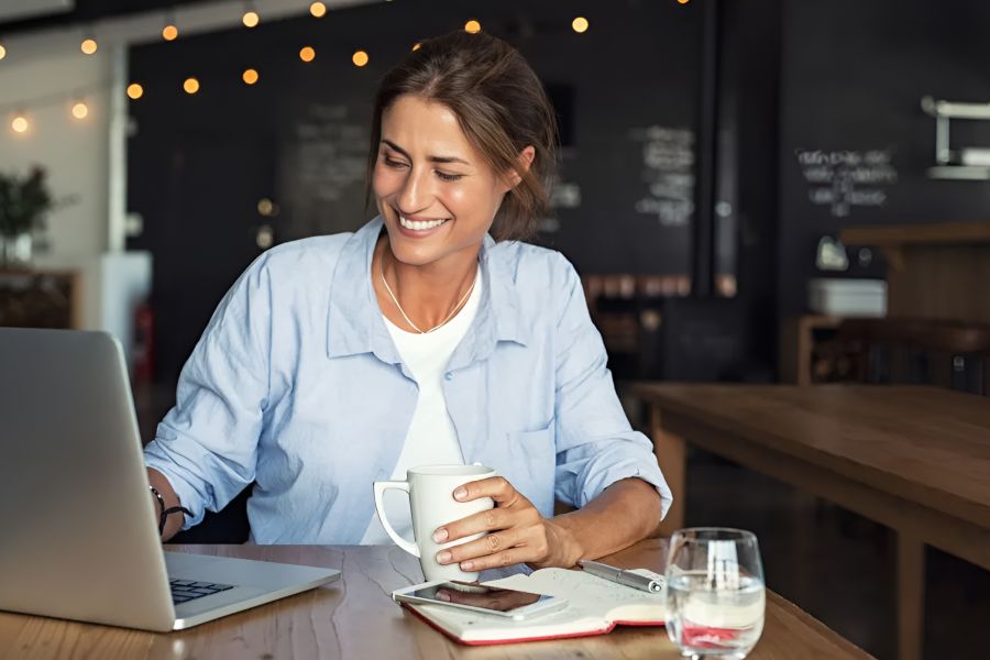 Infinity at Centerville Crossing Smiling woman works on a laptop in a cafe, holding a mug, with a notebook and glass of water nearby.